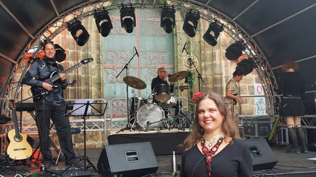 Alan Reynolds, Martin, Lady of Glencoe und Missy K. arbeiten auf einer Veranstaltungsbühne mit halbrundem Dach. Lucinde steht im Vordergrund vor der Bühne und blickt lächelnd in die Kamera. Im Hintergrund ein großes Eingangsportal in ein Sandsteingebäude.